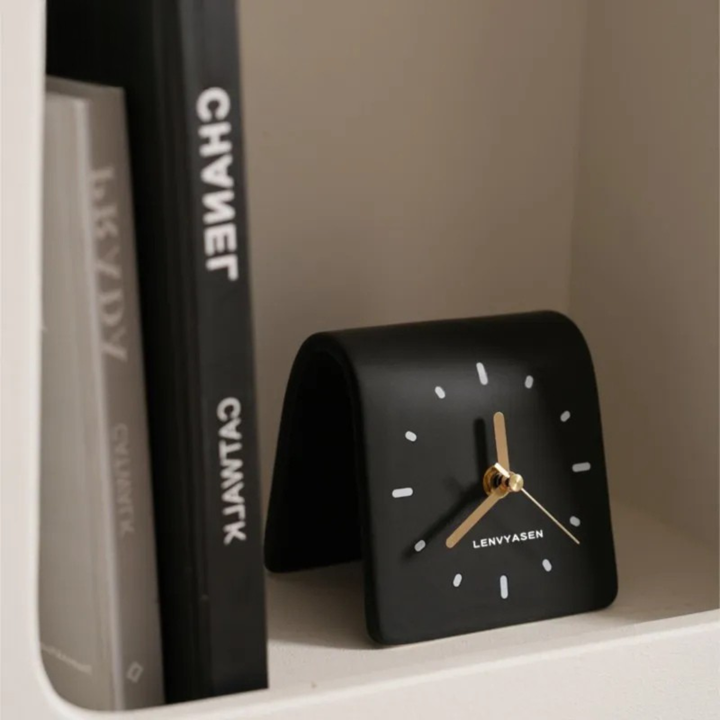 A black leather desk clock with gold hands sitting on a shelf next to a black and white “CHANEL” book.
