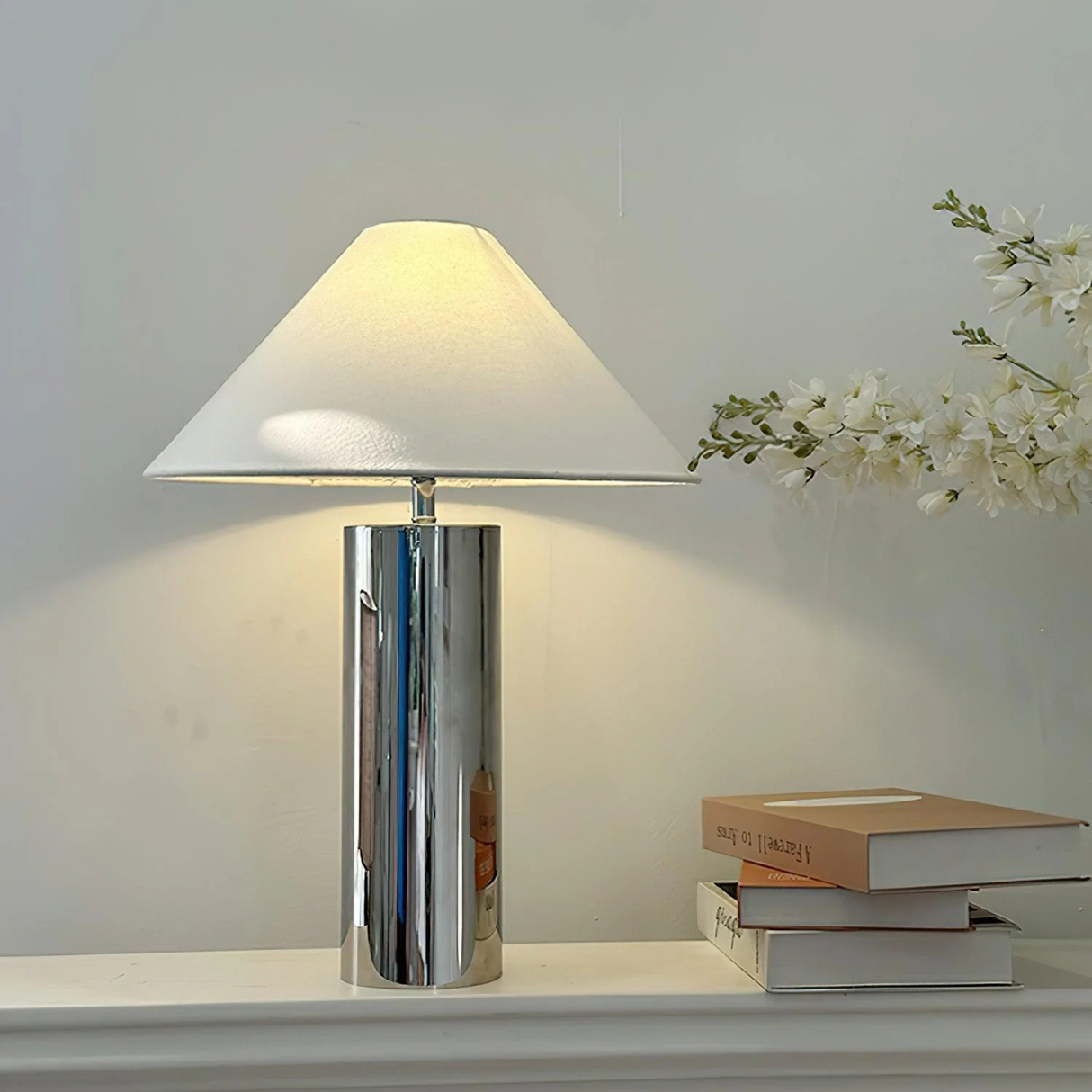 chrome table lamp with warm light beside stacked books and white flowers

