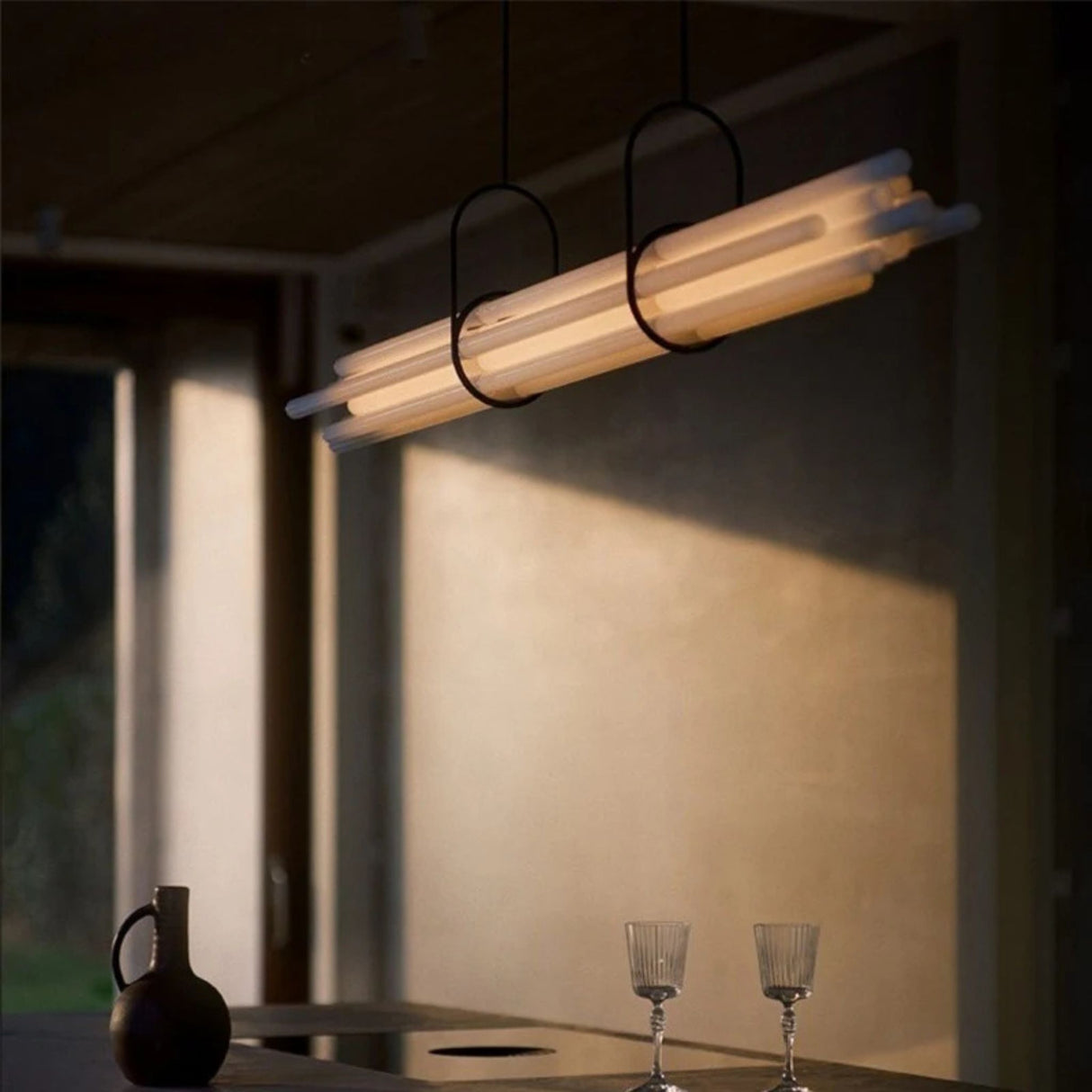 A linear modern pendant light with a frosted diffuser glowing warmly above a minimalist dining table in a softly lit room.
