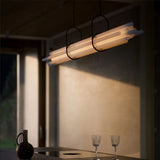A linear modern pendant light with a frosted diffuser glowing warmly above a minimalist dining table in a softly lit room.