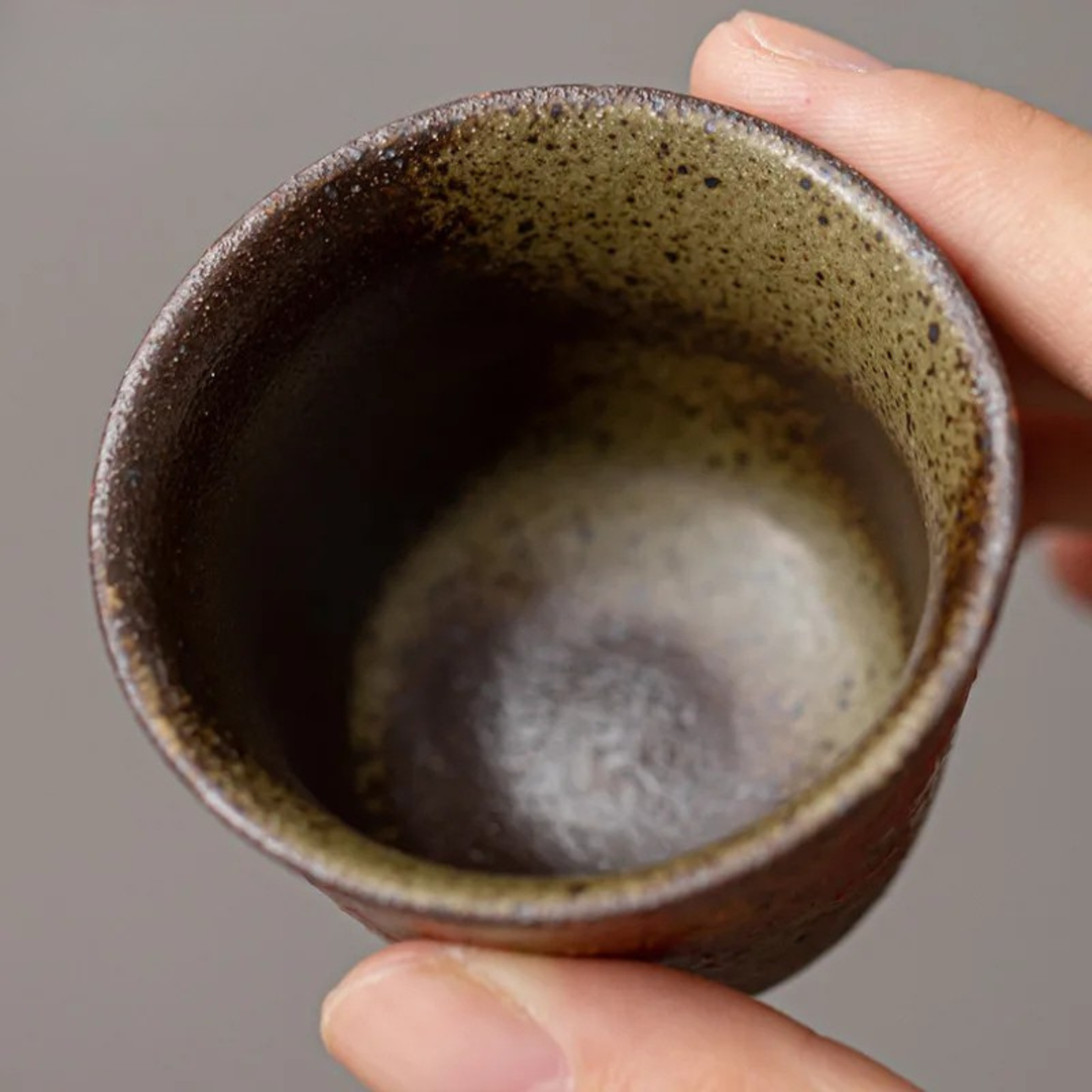 Close-up of a hand holding a small red ceramic tea cup, showing the speckled green interior.