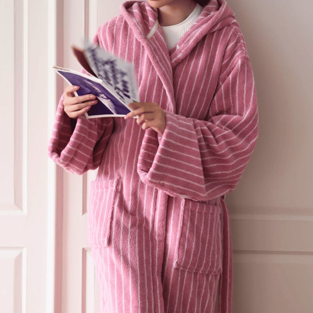 Person wearing a pink robe reading a magazine against a white wall.