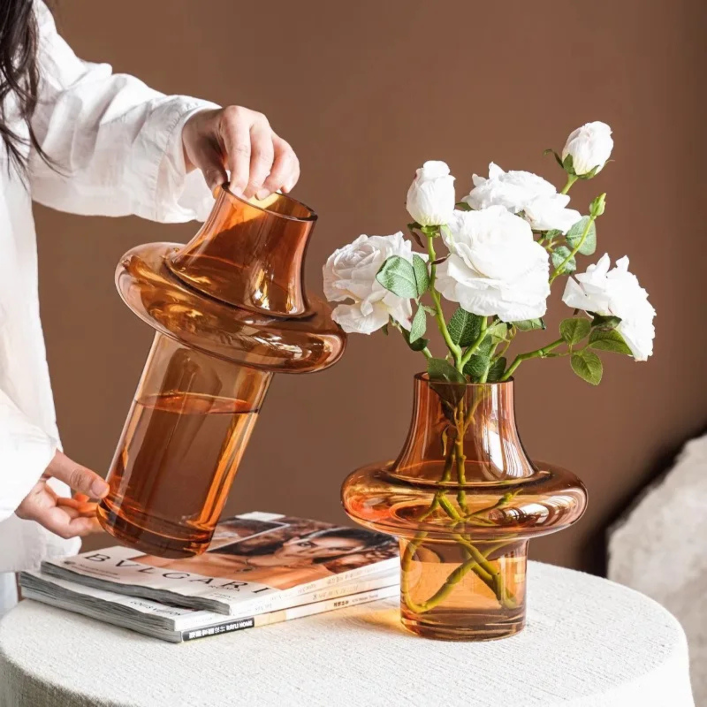 Person holding a tall amber glass vase while another identical vase filled with white flowers sits on a stack of magazines.