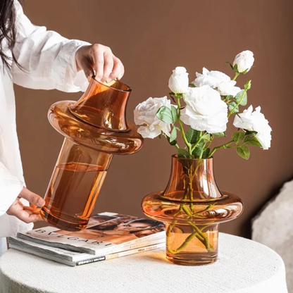 Person holding a tall amber glass vase while another identical vase filled with white flowers sits on a stack of magazines.