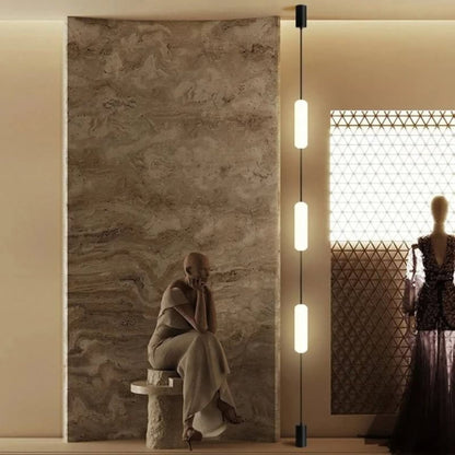 A slim vertical tube pendant light illuminating a textured beige wall near a walk-in shower area.