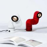 A close-up of a red and white desk lamp, positioned side by side, with the light directed downward onto an open magazine.