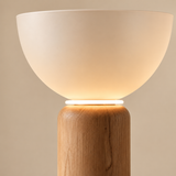 A modern table lamp with a warm white frosted bowl shade and a natural light wood cylindrical base, shown against a neutral beige background.