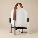 A modern table lamp with a glossy white oval shade, a brown leather top handle, and a polished silver metal stand, set against a solid beige background.