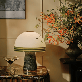 Table lamp with a green base and white shade on a marble table, next to a vase of flowers.