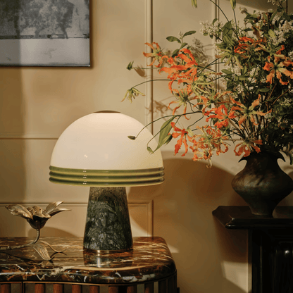Table lamp with a green base and white shade on a marble table, next to a vase of flowers.