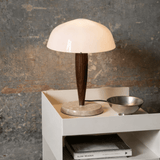 Table lamp with wooden base and white shade on a white surface against a textured wall.