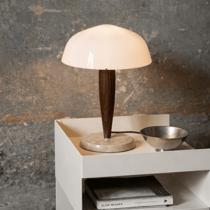 Table lamp with wooden base and white shade on a white surface against a textured wall.