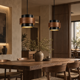 Two modern cylindrical pendant lights with black and warm brown wood-grain shades hang over a rustic wooden dining table in a styled interior room.