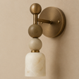 A modern wall sconce with a round brass backplate, a vertical brass stem, stacked matte brass and white marble spheres, and a white marble cylindrical shade, mounted on a beige wall.
