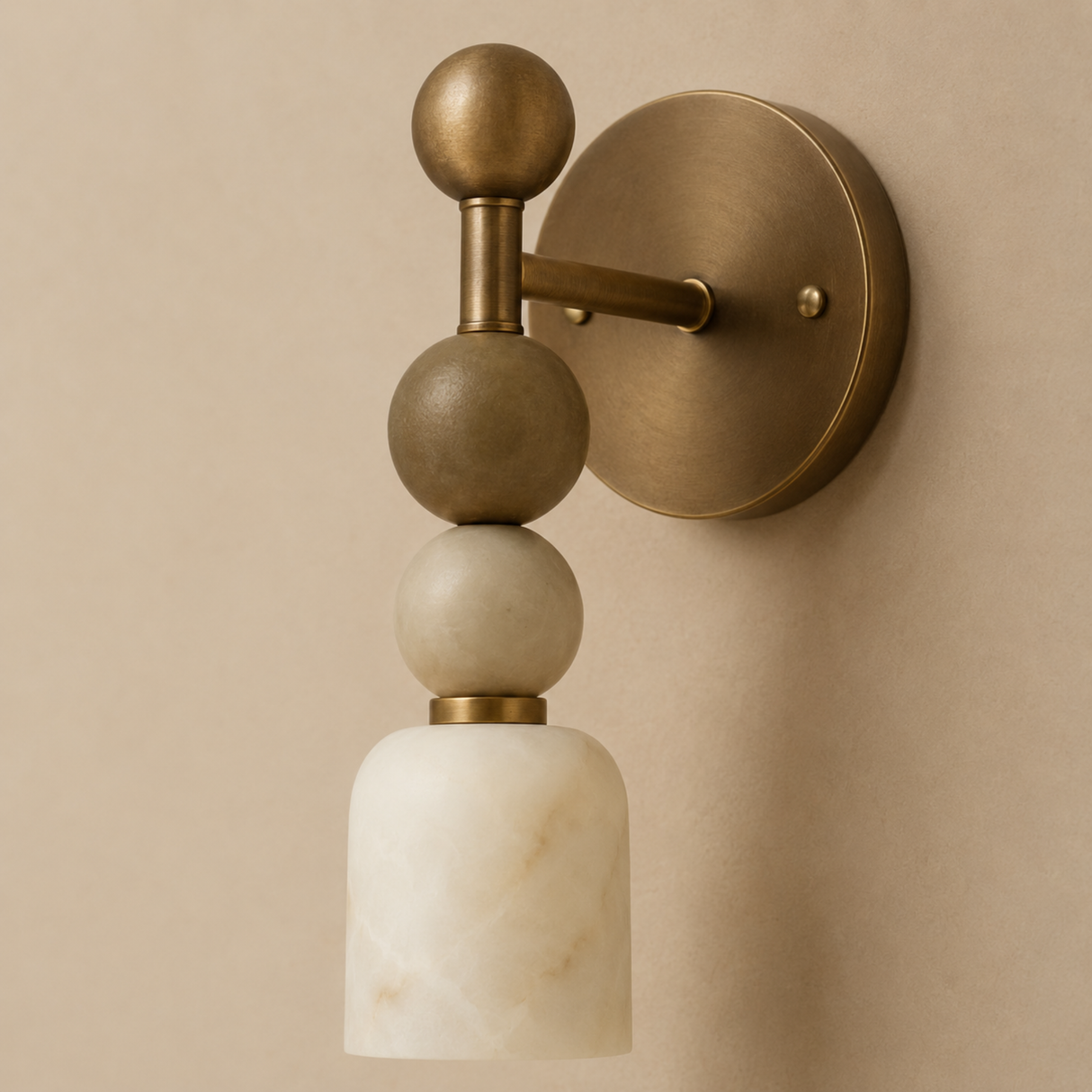 A modern wall sconce with a round brass backplate, a vertical brass stem, stacked matte brass and white marble spheres, and a white marble cylindrical shade, mounted on a beige wall.