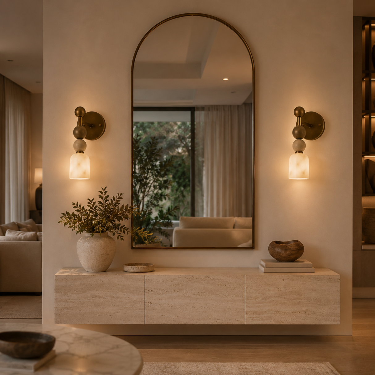 A large arched wall mirror with a bronze frame hangs above a light wood console in a modern interior. Two matching wall sconces glow on either side, and the mirror reflects a softly lit living room.