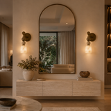 A large arched wall mirror with a bronze frame hangs above a light wood console in a modern interior. Two matching wall sconces glow on either side, and the mirror reflects a softly lit living room.