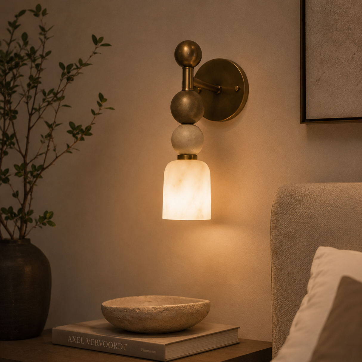 A modern wall-mounted sconce with a warm brass finish and stacked spherical shades, glowing with a warm white light in a bedroom setting.