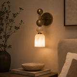 A modern wall-mounted sconce with a warm brass finish and stacked spherical shades, glowing with a warm white light in a bedroom setting.