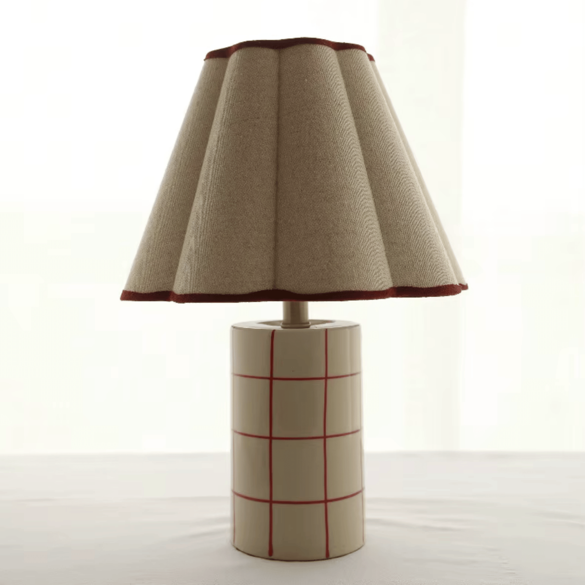 Table lamp with a textured beige shade and a cylindrical base with red grid pattern on a white background