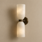 A modern two-light wall sconce with frosted white glass cylindrical shades and a brushed bronze metal mount, shown lit against a warm beige wall.