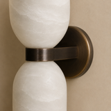 A close-up of a wall-mounted two-light fixture with white textured glass shades and a brushed bronze metal band and backplate, set against a beige wall.