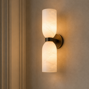 A modern wall-mounted sconce with two cylindrical frosted glass shades and a dark metal mount, shown lit against a beige wall.