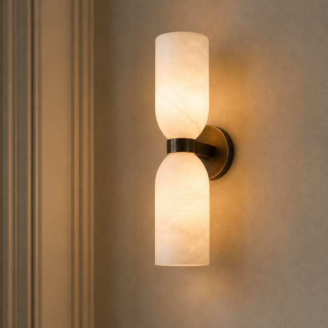 A modern wall-mounted sconce with two cylindrical frosted glass shades and a dark metal mount, shown lit against a beige wall.