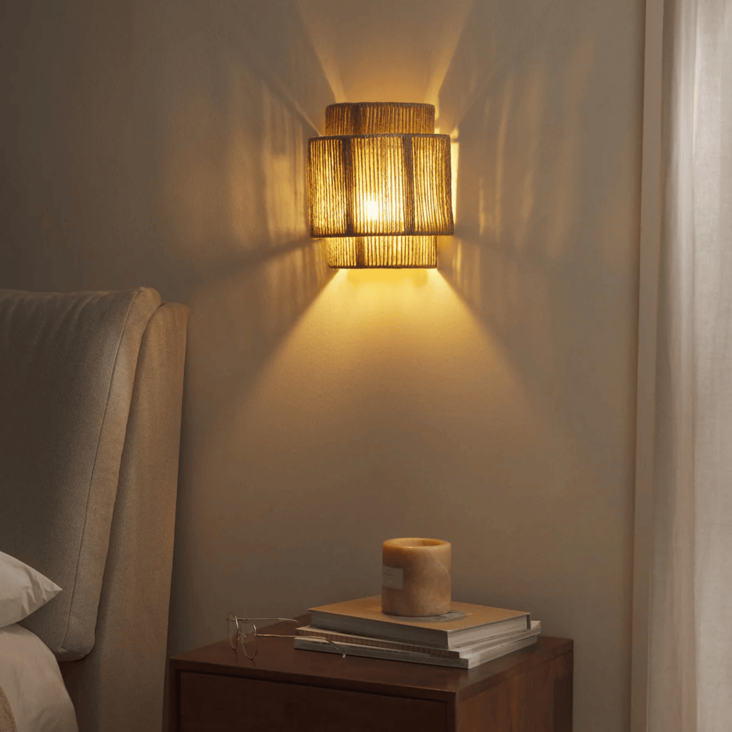 Woven wall light fixture casting a warm glow on a wall next to a nightstand with books and a candle.