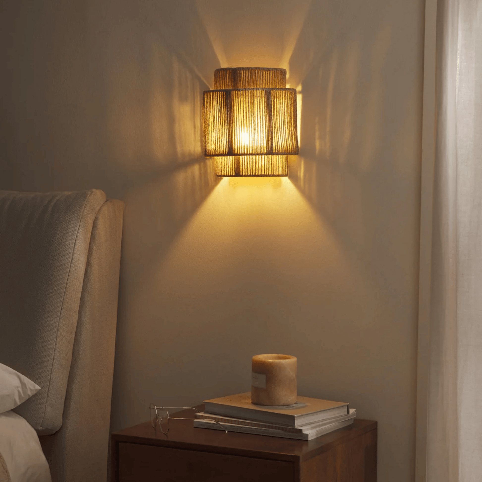 Woven wall light fixture casting a warm glow on a wall next to a nightstand with books and a candle.