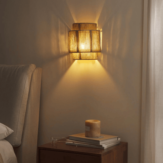 Woven wall light fixture casting a warm glow on a wall next to a nightstand with books and a candle.