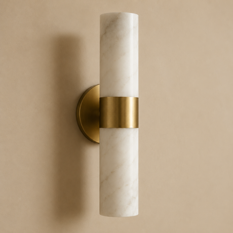 A modern wall-mounted light fixture with a cylindrical white marble shade and a brushed gold metal band and wall mount, shown against a warm beige background.
