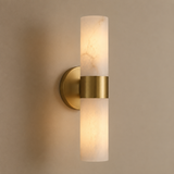 A modern cylindrical wall sconce with a brushed gold metal band and a frosted white marble-look glass shade, shown lit against a warm beige wall.