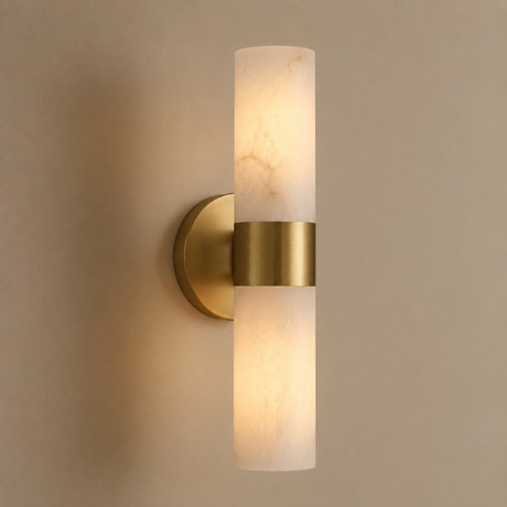 A modern cylindrical wall sconce with a brushed gold metal band and a frosted white marble-look glass shade, shown lit against a warm beige wall.
