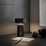 Dark table lamp on a surface with a pen and paper in a minimalistic setting.