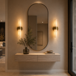 An oval wall mirror with a thin black frame hangs above a light wood floating vanity in a warm, modern interior. Two matching vertical wall sconces with warm glowing glass shades flank the mirror, and a dark glass vase with dried greenery sits on the console.