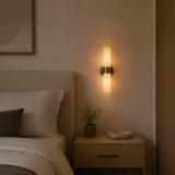 A modern wall-mounted sconce with a vertical frosted glass shade and a brushed brass or bronze metal base, illuminated in a warm light next to a bed in a minimalist bedroom setting.
