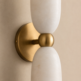 A modern wall-mounted light fixture with two elongated white marble shades and a brushed brass center joint and circular backplate, shown against a warm beige wall.