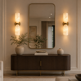 A styled interior scene showing a dark wood console table with gold legs, a large wall mirror, two illuminated wall sconces, and decorative vases on top, set against a beige paneled wall.
