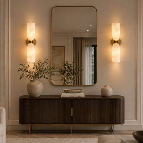 A styled interior scene showing a dark wood console table with gold legs, a large wall mirror, two illuminated wall sconces, and decorative vases on top, set against a beige paneled wall.