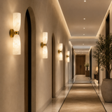 A modern double-light wall sconce with a brushed brass fixture and two glowing white textured glass shades, mounted on a beige wall in a long, elegantly lit hallway.