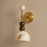 A wall-mounted sconce with a round gold brass backplate, a vertical gold stem, a white marble sphere finial, two stacked dark brown marble beads, and a white marble dome shade with brown veining, shown against a warm beige wall.