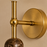 Close-up of a wall-mounted light fixture with a brushed brass circular backplate, horizontal arm, and vertical stem, featuring a dark brown marble globe shade against a warm beige wall.