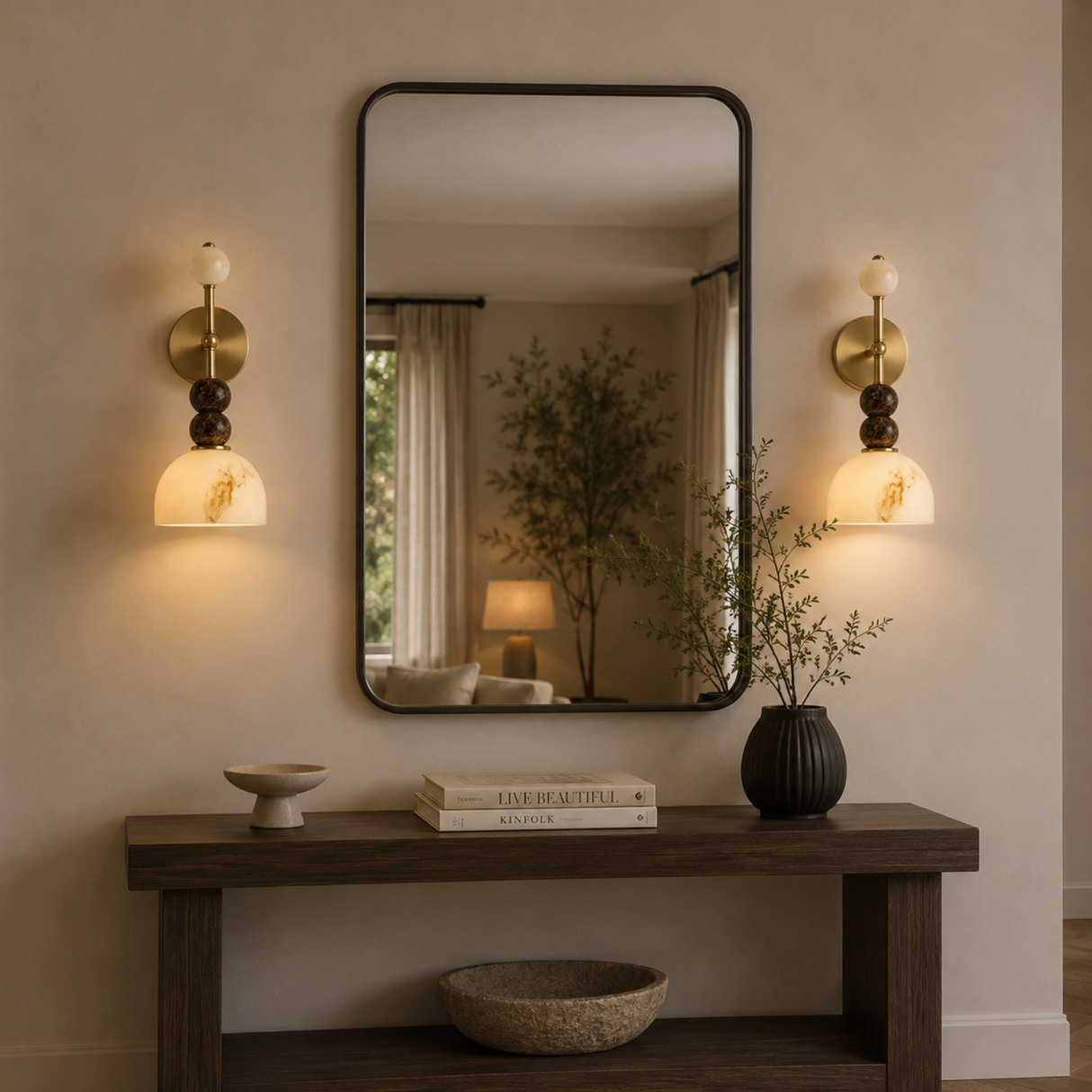 A large rectangular wall mirror with a thin black frame hangs above a dark wood console table in a warm, modern interior. Flanking the mirror are two matching wall sconces with warm glowing glass shades.