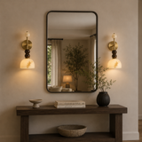 A large rectangular wall mirror with a thin black frame hangs above a dark wood console table in a warm, modern interior. Flanking the mirror are two matching wall sconces with warm glowing glass shades.
