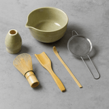 Set of tea-making tools including a green bowl, small vase, whisk, spoon, and strainer on a gray surface.