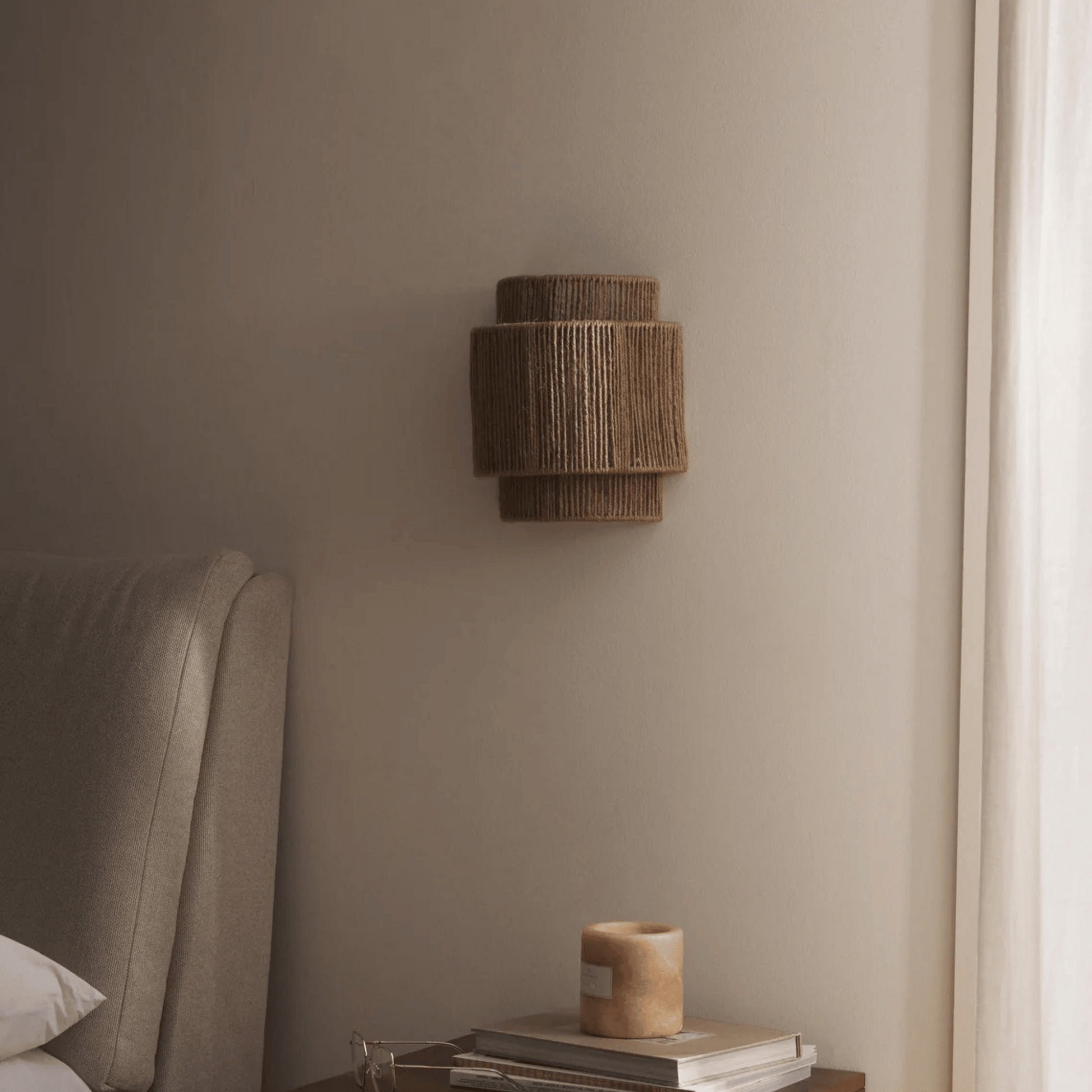 Woven wall light fixture on a neutral wall next to a bed with a lamp and books.