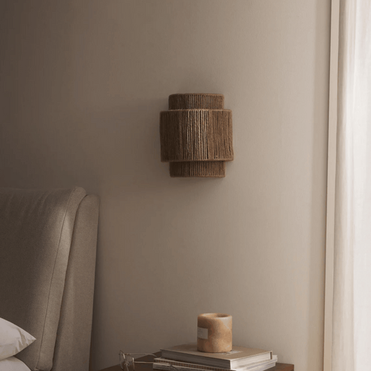 Woven wall light fixture on a neutral wall next to a bed with a lamp and books.