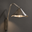 A modern wall-mounted sconce with a brushed metal finish, featuring a curved gooseneck arm and a flared shade that casts a warm light against a dark gray wall.