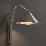 A modern wall-mounted sconce with a brushed metal finish, featuring a curved gooseneck arm and a flared shade that casts a warm light against a dark gray wall.